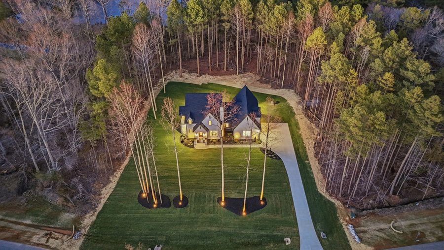 February 2026 dusk aerial of a completed home at Southern Pine Lake with the drive and young trees uplit.