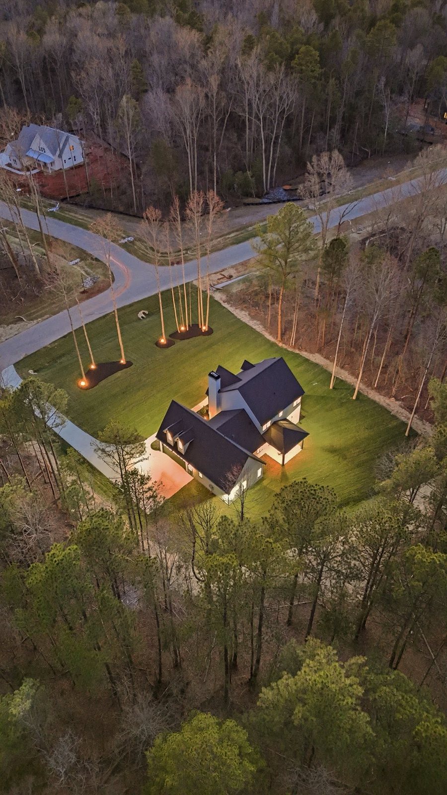 Dusk aerial of a completed home at Southern Pine Lake, uplit on its cleared homesite with pine and hardwood around it.