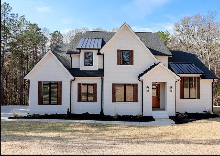 The first completed custom home in the community, white brick with dark metal roof accents and stained wood shutters.