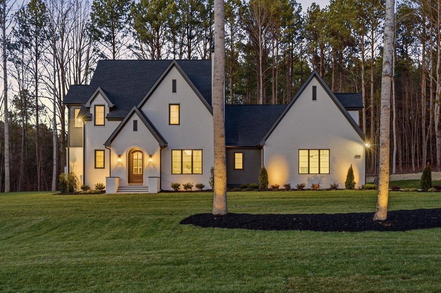 White brick custom home at dusk, uplit against the treeline, with an arched wood entry door.